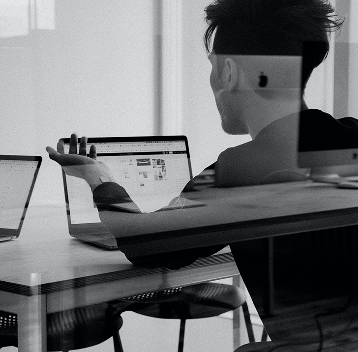 Grayscale image of a man looking at something on his laptop; his back is facing the camera